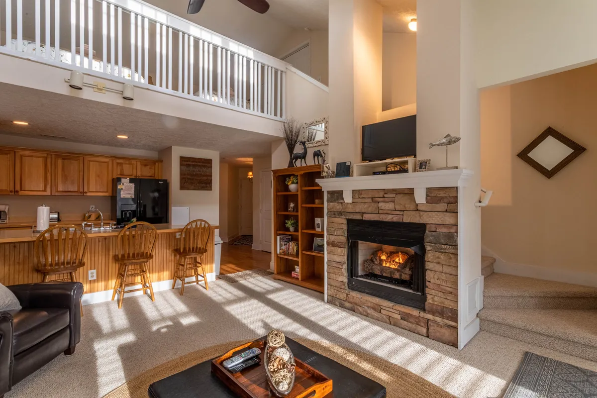 A cozy open-concept living area featuring a plush seating arrangement centered around a warm gas log fireplace. The space flows seamlessly into a modern kitchen and loft area, with sunlight streaming through large windows, creating a welcoming and comfortable atmosphere ideal for family gatherings and relaxation.