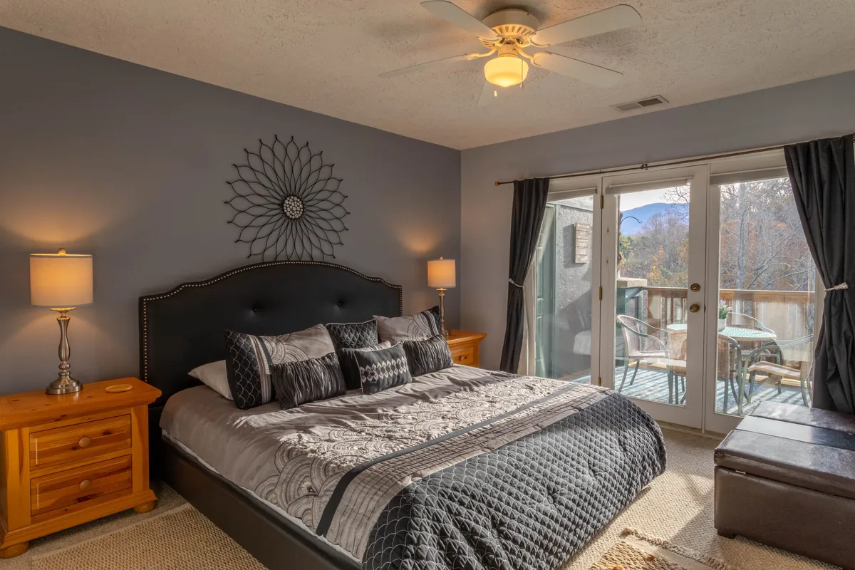 A serene master bedroom featuring a king-sized bed with plush bedding. Decorative wall art adds character, while a sliding glass door opens directly onto a private balcony or deck, allowing natural light to flood the room. The tranquil setting is enhanced by soft lighting and tasteful decor.