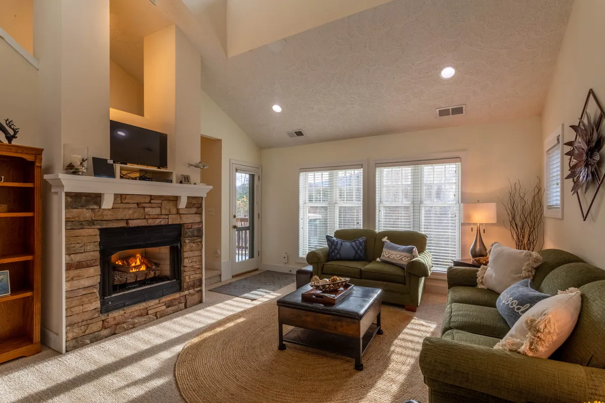 The image showcases a warm, inviting living area with plush couches gathered around a stone fireplace, large windows framing serene mountain views, and a spacious coffee table perfect for family activities. Sunlight pours in, creating a bright yet cozy atmosphere that invites relaxation and togetherness.