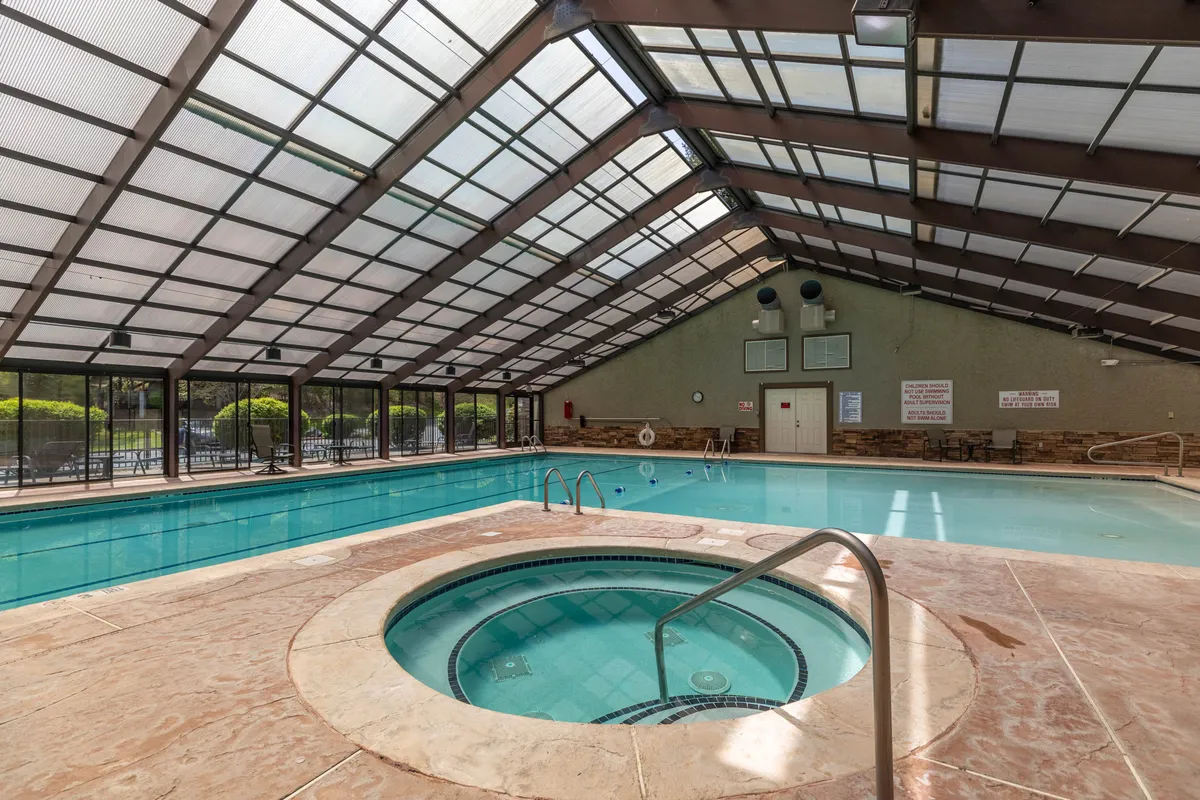 This image showcases a spacious indoor swimming pool alongside a relaxing hot tub, with large glass windows letting in natural light and a distinctive roof structure overhead. The area is designed for both swimming and unwinding, encouraging social and individual activities.