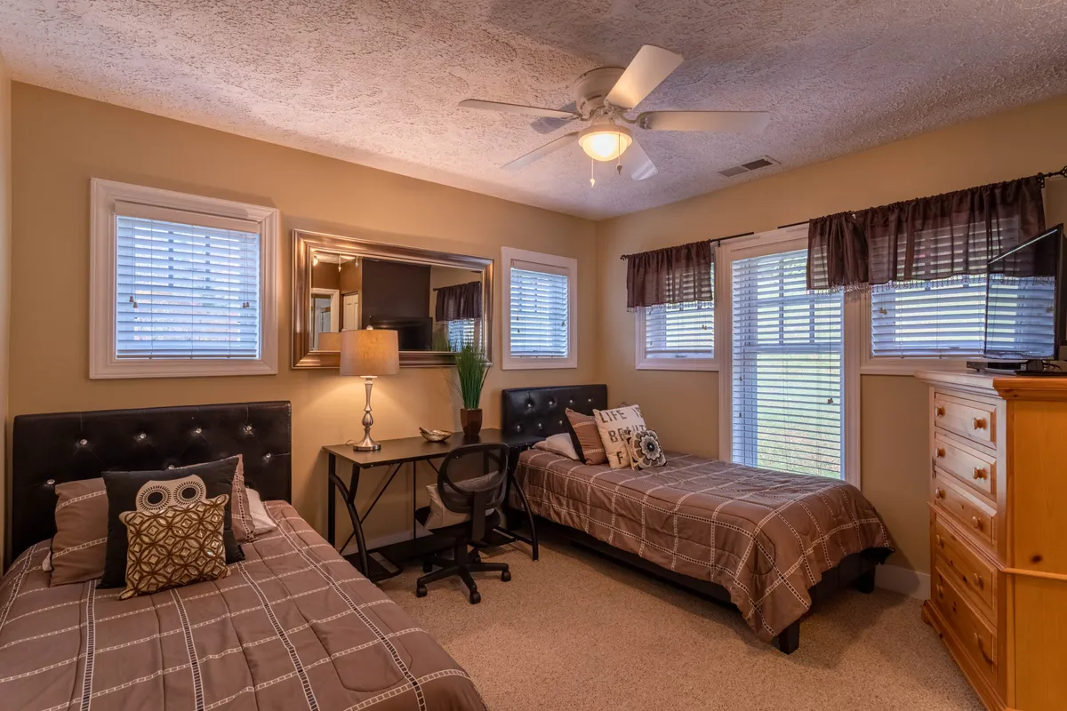 A cozy bedroom with two twin beds, a desk with a chair, a large mirror, and a lamp. The arrangement is practical and inviting, making it ideal for kids or friends sharing the space. Personal touches like the dresser and soft lighting add warmth.