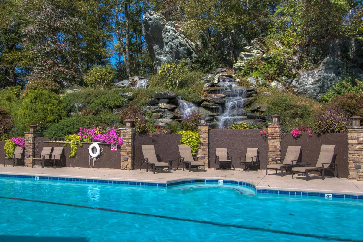 An inviting outdoor pool area featuring a sparkling swimming pool, rocky waterfall feature, lounge chairs, and vibrant flowering plants. The serene setting offers mountain views and a resort-style atmosphere ideal for relaxing or socializing.