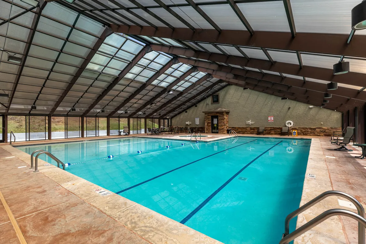 A bright indoor clubhouse featuring an inviting swimming pool surrounded by large glass windows, wooden beams, and comfortable seating areas, offering both recreational and gathering spaces for guests.