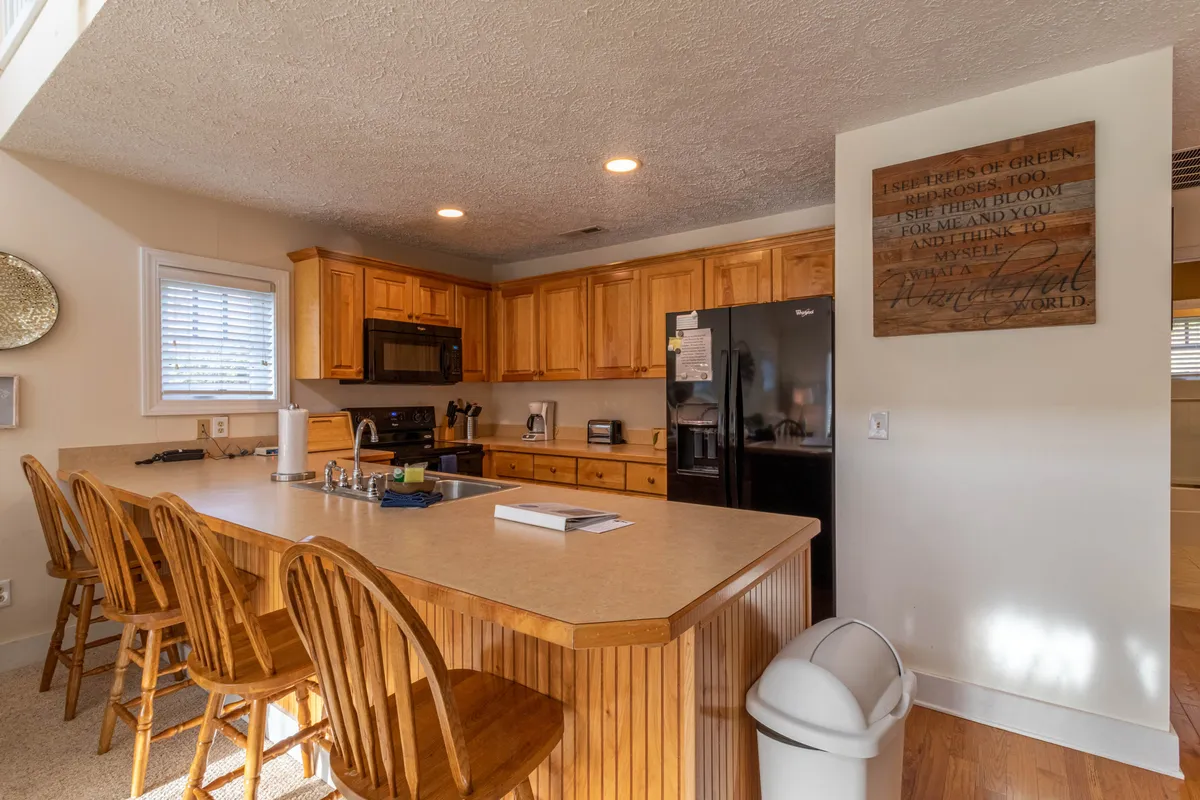 This image depicts a warm, welcoming kitchen with a prominent island, wooden cabinetry, a modern black refrigerator, and countertop appliances. The space is bright and inviting, ideal for meal preparation and gathering with family.